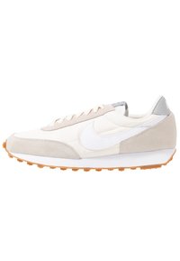 Ljust beige atletiska skor med en slät textilöverdela, mockadetaljer, vit swoosh-logotyp och en gummisula med orangea greppknoppar.