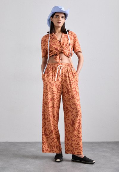 Orange floral-patterned cropped shirt with knotted front, matching wide-leg pants, and black flat shoes. Blue bucket hat with chin straps.