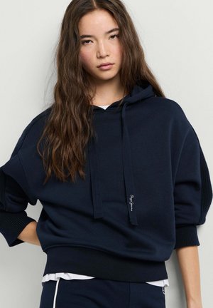Jonge vrouw met lang bruin haar, gekleed in een losse donkerblauwe hoodie en bijpassende broek tegen een effen lichte achtergrond.