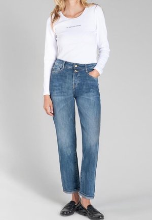 Kvinde iført hvid skjorte med lange ærmer med tekst og blå højtaljede jeans med dobbelte knapper, stående med den ene hånd i lommen.