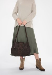 Bruine leren handtas met een gestructureerd voorpaneel, gedragen over een lichtgroene rok en een beige trui, gecombineerd met tan enkellaarsjes.