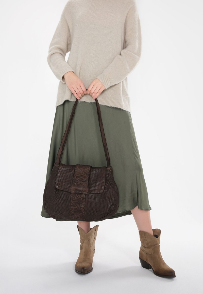 Bruine leren handtas met een gestructureerd voorpaneel, gedragen over een lichtgroene rok en een beige trui, gecombineerd met tan enkellaarsjes.