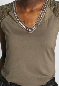 Olive green top with lace shoulder accents, V-neckline, and metallic trim details. Smooth textured fabric with a relaxed fit.