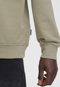 Khaki trui met structuurstof, geribbelde manchetten en een zichtbaar label. Close-up van een hand die het draagt, op een donker kledingstuk.