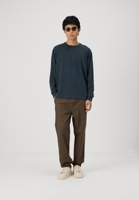 Dark teal long-sleeve shirt paired with brown pants. The shirt has a crew neckline, and the model wears beige sneakers and sunglasses.