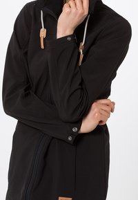 Chaqueta softshell negra con cremallera, capucha ajustable y cierres de madera. Presenta botones a presión y una textura suave.