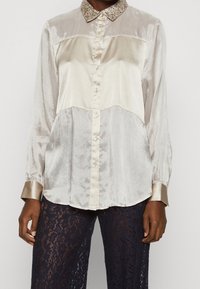 Camisa de botones de seda en color crema con un cuerpo texturizado, cuello beige contrastante y mangas largas. Acabado con un borde de collar con lentejuelas.