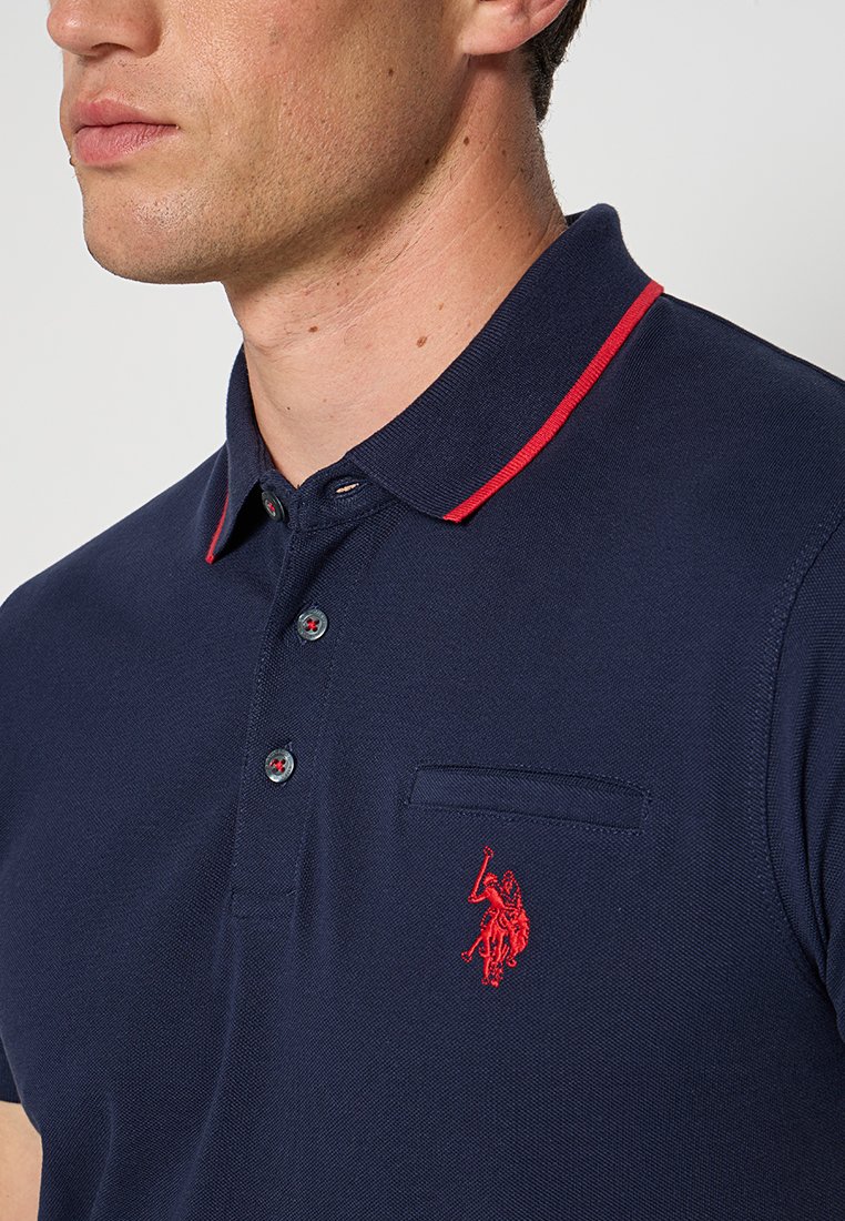 Marineblaues Poloshirt aus strukturiertem Stoff, mit einem roten Kragenstreifen, einer Knopfleiste mit zwei Knöpfen und einem roten gestickten Logo auf der Brusttasche.