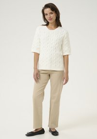 Pull en tricot blanc avec un motif texturé, manches trois-quarts, associé à un pantalon beige et des ballerines noires.