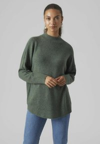 Oversized green knitted sweater with a high neck, ribbed cuffs, and hem, paired with blue jeans. Soft texture and relaxed fit.