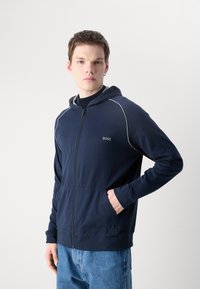 Felpa con zip di colore blu navy realizzata in tessuto morbido, dotata di cappuccio, tasche frontali e piping bianco a contrasto lungo le spalle.