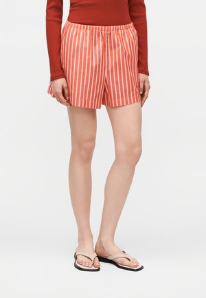 Γυναίκα που φοράει πορτοκαλί και λευκά κάθετα ριγέ σορτς, ένα μακρυμάνικο τοπ σκουριάς και μπεζ σαγιονάρες, στέκεται μπροστά σε λευκό φόντο.