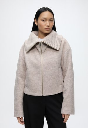 Vrouw met lang zwart haar, wearing een beige jas met ritssluiting en grote geribbelde kraag, en zwarte broek tegen een effen lichte achtergrond.