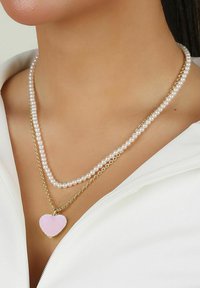 Tredubbelhalsband med två strängar av vita pärlor och en guldkedja, med en rosa hjärtanpendel som en anmärkningsvärd accent.