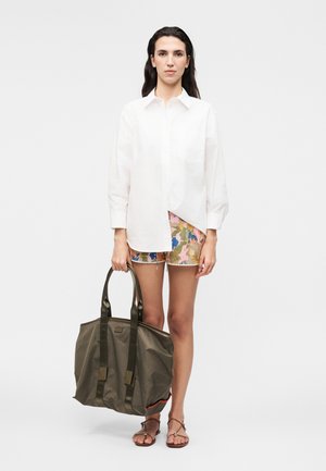 Femme portant une chemise blanche à manches longues, un short à fleurs, des sandales, tenant un grand sac fourre-tout vert olive sur un fond blanc uni.