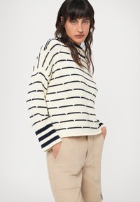 Kreminė megzta palaidinė su tamsiai mėlynomis horizontaliomis juostelėmis ir tekstūruotu raštu. Laisvas fasonas su gumiukais ant rankovių ir apvaliu iškirpimu.