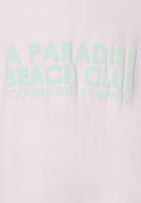 Vaaleanpunainen t-paita, jossa on rohkeaa mintunvihreää tekstiä, joka lukee "PARADISE BEACH CLUB" ja "PERUSTETTU 2017 @PEQUS:n TOIMESTA". Pehmeä kangas.