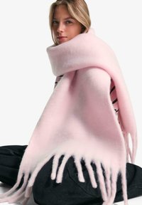 Écharpe légère rose pâle en matériau doux, avec des bords à franges et un design simple, drapée sur une personne assise par terre.