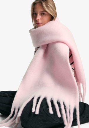 Fluffy lichtroze sjaal van zacht materiaal, met franjes aan de randen en een eenvoudig ontwerp, over een persoon die op de grond zit.