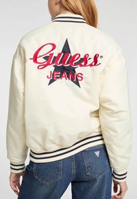 Witte bomberjack met contrasterend zwart en rood geborduurd "Guess Jeans" logo en een sterontwerp. Voorzien van gestreepte geribde manchetten en zoom.