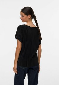 Donna con capelli intrecciati che indossa una maglietta nera a maniche corte plissettata e jeans blu scuro, rivolta di lato su uno sfondo bianco.