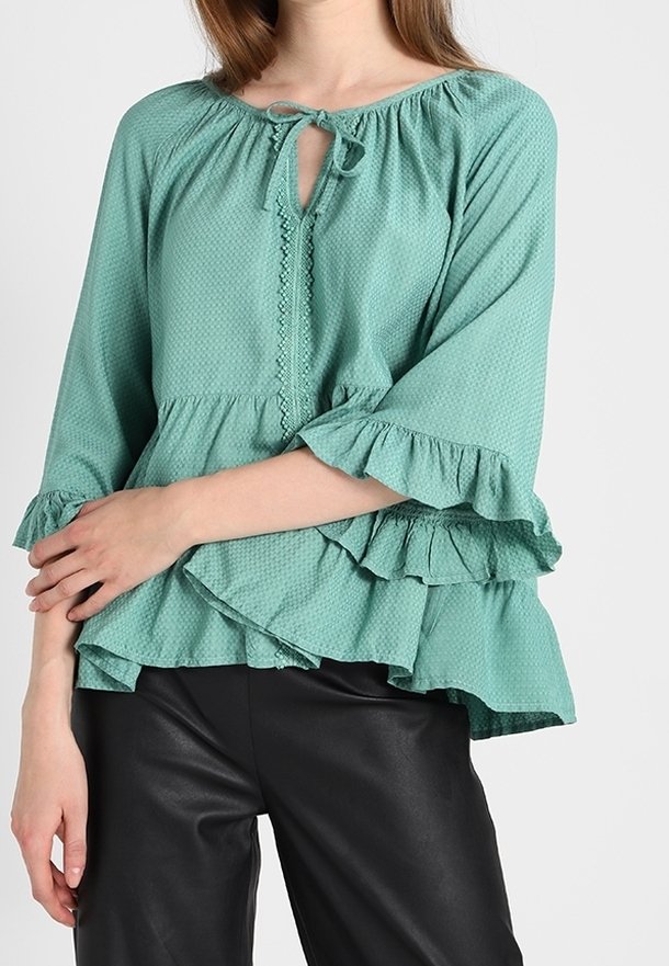 Femme portant un chemisier vert texturé à manches longues avec des manches et un ourlet volantés, assorti à un pantalon en cuir noir.