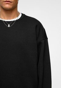 Zwarte sweatshirt met ronde hals, gemaakt van zachte stof. Heeft ontspannen mouwen en een gelaagd detail van een witte shirt eronder.