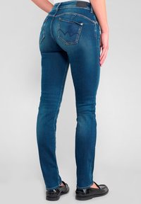Jean en denim bleu skinny avec une taille mi-haute, poches arrière avec un design subtil et une texture lisse. Chaussures plates noires visibles.