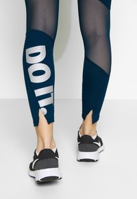 Navy leggings with mesh panels, featuring a prominent silver "DOTT." logo, and side slits at the ankles. Worn with black athletic shoes.