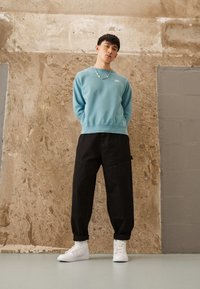 Lichtblauwe sweatshirt met geribde zoom, zwarte baggy broek en witte sneakers, tegen een gestructureerde, neutrale muur. Zilveren kettinghalsketting.