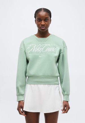 Νεαρή γυναίκα που φοράει φούτερ Nike σε μέντα πράσινο και λευκή φούστα τένις, στέκεται μπροστά σε ένα απλό φωτεινό φόντο.