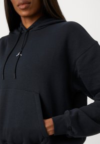 Sweatshirt à capuche noire en tissu doux, avec une poche frontale, un cordon de serrage et un détail logo subtil sur la poitrine.