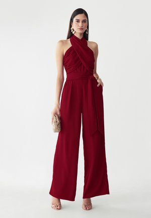 Donna che indossa una tuta bordeaux senza maniche con scollo halter e gambe larghe, tiene una piccola pochette testurizzata e indossa orecchini e braccialetti dorati.