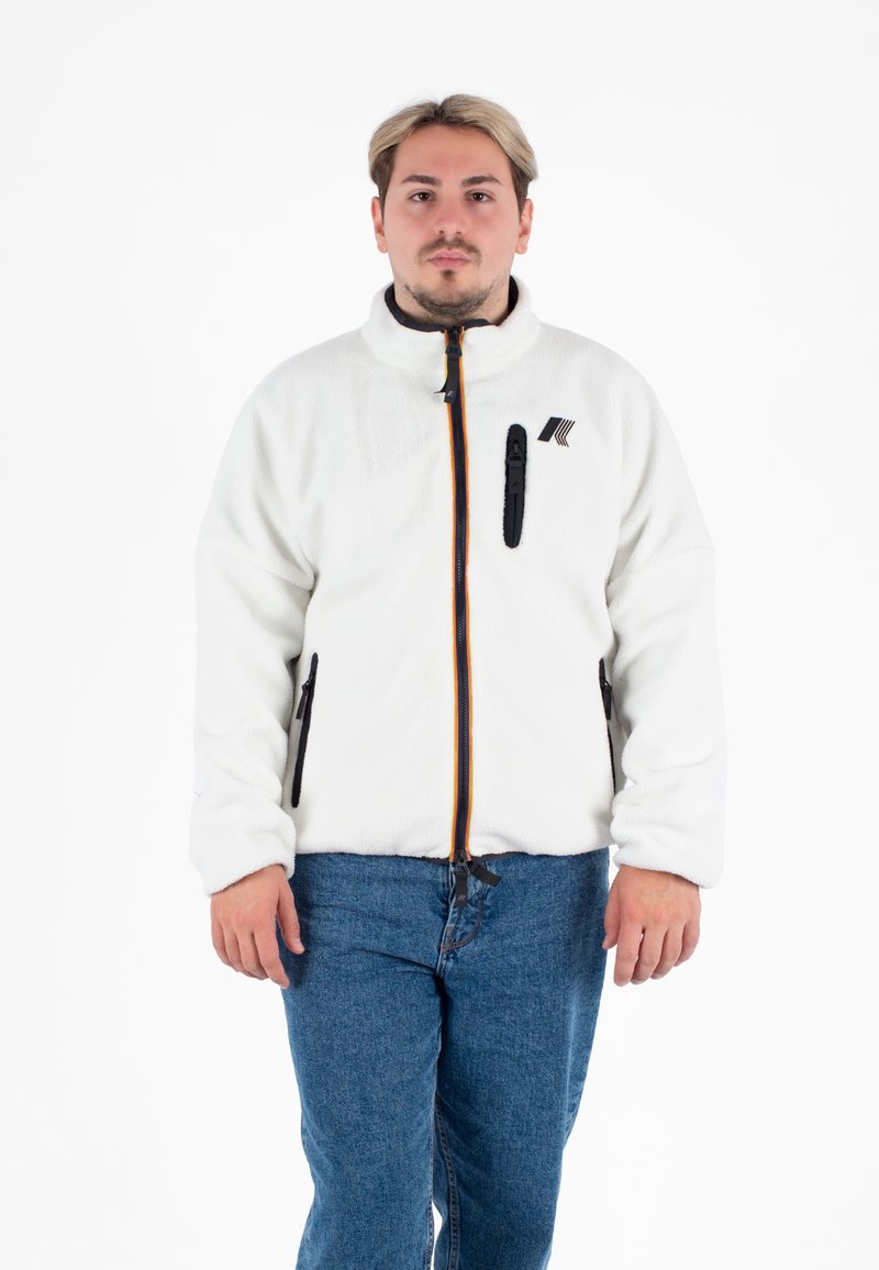 Biała polarowa kurtka z stójką, z czarnymi zamkami, pomarańczowym akcentem i kieszenią na piersi. Nosi się na niebieskich dżinsach.