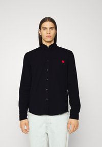 Zwarte langslang shirt met knoop-kraag, voorzien van een borstzak en een klein rood logo. Gemaakt van zachte stof, met een relaxte pasvorm en een rond afgeknipte zoom.