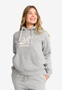 Leende kvinna med blont hår iklädd en ljusgrå hoodie och matchande sweatpants, med händerna i fickorna, tittande uppåt och åt sidan.