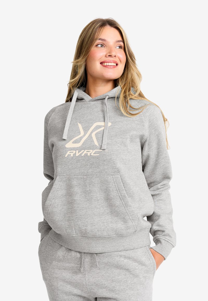 Leende kvinna med blont hår iklädd en ljusgrå hoodie och matchande sweatpants, med händerna i fickorna, tittande uppåt och åt sidan.