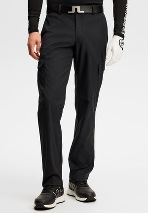 Mand ifører sig sorte cargobukser, sort bælte med sølvspænde, sorte sneakers med hvide såler og en hvid golfhandske på venstre hånd.