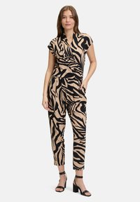Tuta color tan con strisce zebrate nere, maniche corte, design avvolgente, tasche laterali e gambe affusolate. Abbinato a sandali neri con tacco alto.
