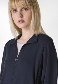 Giacca zip-up navy con colletto alto, realizzata in tessuto testurizzato, presenta una zip argentata ed è sovrapposta a un top rosa chiaro.