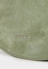 Žalia zomšinė rankinė su lygiu paviršiumi. Su sidabriniu "Cocconelle" logotipu ir kontrastinėmis siūlėmis krašte. Apvalios formos.