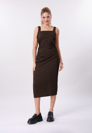 Kvinde med blondt hår iført en ærmeløs, mørkebrun midi-kjole og sorte platåsko, stående mod en ensfarvet lys baggrund.