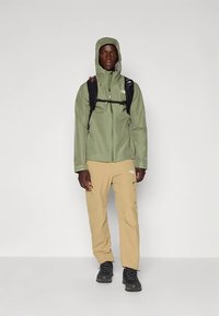 Green waterproof jacket with a hood, beige cargo pants, and black hiking boots. Features a backpack with straps over the jacket.