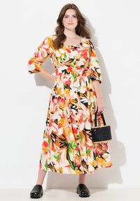 Maxikleid mit floralen Mustern in Orange, Pink, Grün und Schwarz, Rundhalsausschnitt, Knopfleiste und einem Bindegürtel. Trägt eine schwarze Tasche.