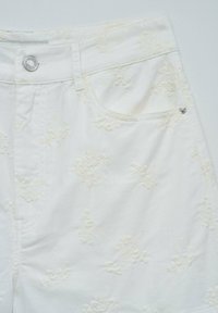 Pantalon en denim blanc avec des motifs floraux en relief, fermeture à bouton en métal, passants de ceinture et une poche avant avec un détail de rivet.