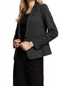 Kvinna som bär en mörkgrå texturerad kavaj över en svart topp och svarta byxor, accessoriserad med guldringar, armband och en kedjehalsband.