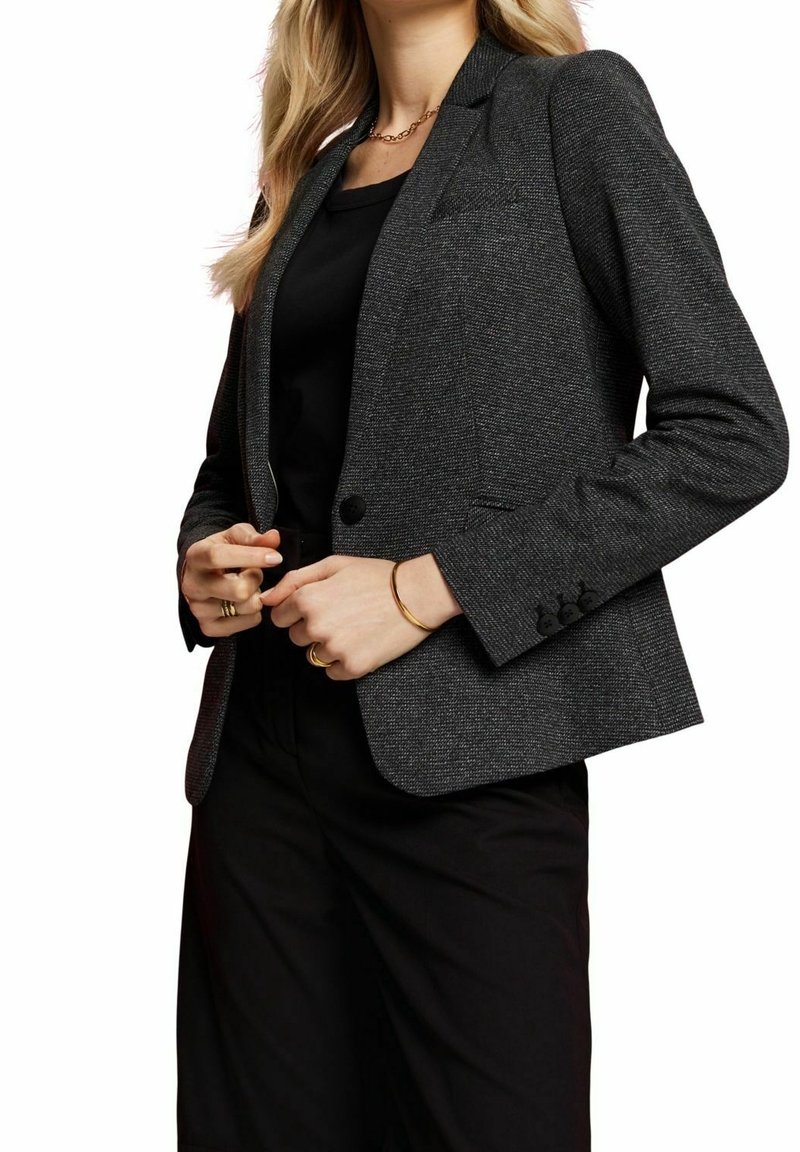 Kvinna som bär en mörkgrå texturerad kavaj över en svart topp och svarta byxor, accessoriserad med guldringar, armband och en kedjehalsband.