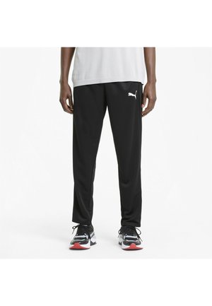 Person trägt schwarze Puma-Trainingshose, weißes T-Shirt und schwarz-weiße Sneakers mit roten Sohlen, steht vor weißem Hintergrund.