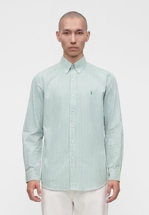 Homme portant une chemise boutonnée à rayures verticales vertes et blanches avec un petit logo brodé sur la poitrine, debout devant un fond uni.