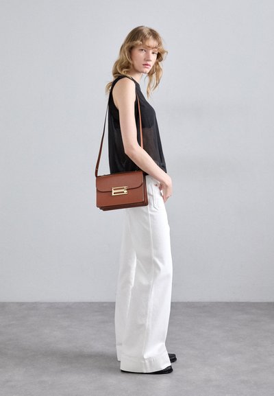 Una persona se encuentra en un entorno minimalista, vestida con una camiseta negra sin mangas y pantalones blancos, sosteniendo un bolso marrón de hombro con un broche dorado.